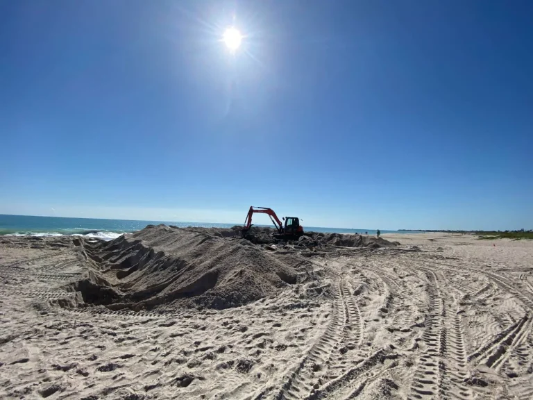 Excavator reshaping a beach dune under bright sun