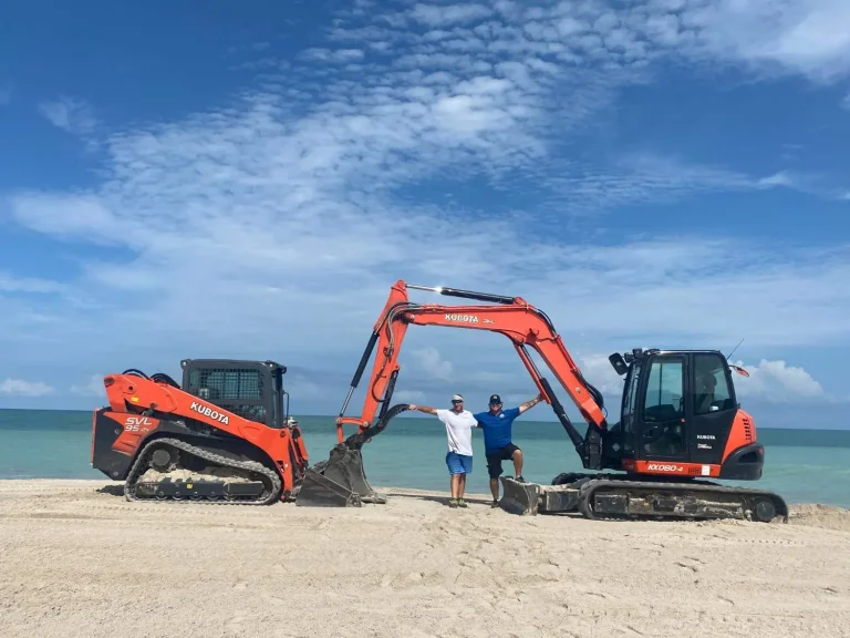 Kubota track loader and excavator on a beach with two crew members