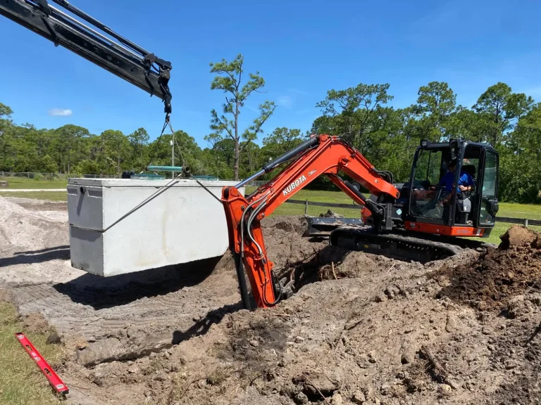 Excavator guiding a septic tank lifted by a crane truck